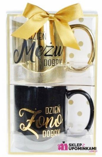Zestaw dwóch kubków w przezroczystym opakowaniu prezentowym: złoty kubek z napisem 'Dzień Męża Dobry' i czarny kubek z napisem 'Dzień Żony Dobry', przewiązane złotą kokardą.