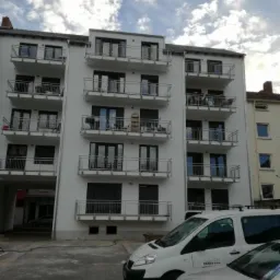 Nowoczesny, biały budynek wielorodzinny z balkonami zabezpieczonymi metalowymi barierkami, częściowo z opuszczonymi roletami zewnętrznymi, widoczny od dołu, z zaparkowanymi samochodami dostawczymi...