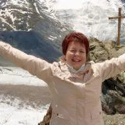 Uśmiechnięta kobieta w beżowej kurtce na tle górskiego krajobrazu z krzyżem. Radosna poza z rozłożonymi ramionami. Jasne, naturalne oświetlenie.