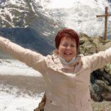 Uśmiechnięta kobieta w beżowej kurtce na tle górskiego krajobrazu z krzyżem. Radosna poza z rozłożonymi ramionami. Jasne, naturalne oświetlenie.