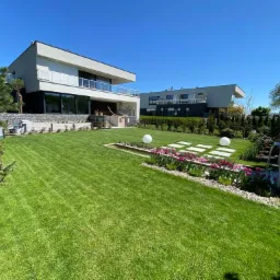 Perfekcyjnie utrzymany trawnik przed nowoczesnym domem z minimalistycznym ogrodem, fontanną i dwoma dużymi kulami oświetleniowymi.