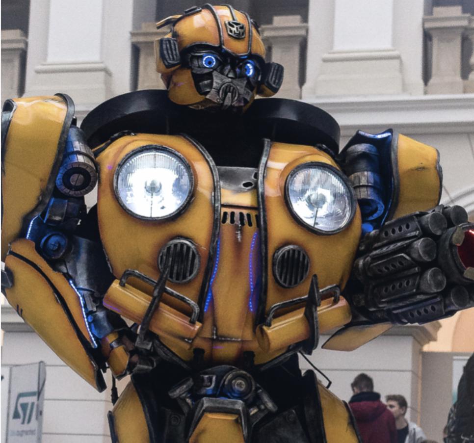 Żółto-czarny kostium robota Bumblebee z niebieskimi detalami, z przodu reflektory, w tle fragment budynku.