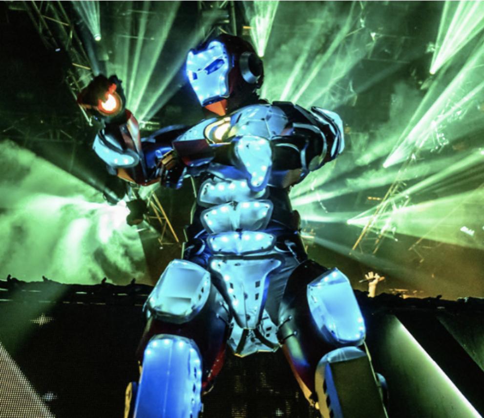 Osoba w kostiumie robota z podświetleniem LED na scenie podczas wydarzenia, oświetlona zielonymi reflektorami.