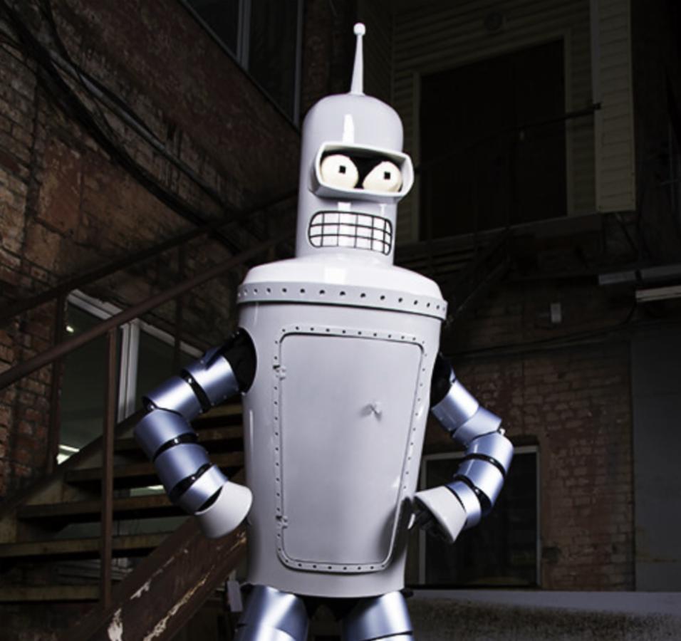 Duży, srebrny robot o wyglądzie znanym z kreskówki, z rękami założonymi na biodra, stoi na tle ceglanej ściany i schodów.