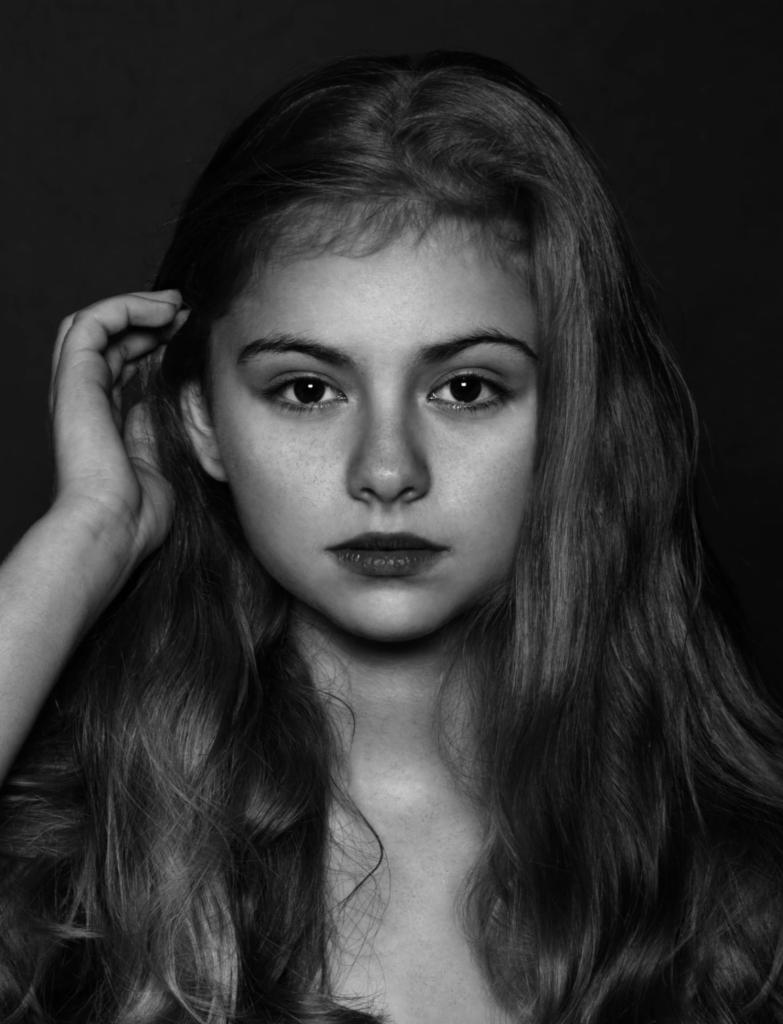 Czarno-biały portret młodej kobiety z długimi, falującymi włosami, patrzącej prosto w obiektyw z poważnym wyrazem twarzy, ręka uniesiona do włosów po lewej stronie kadru.