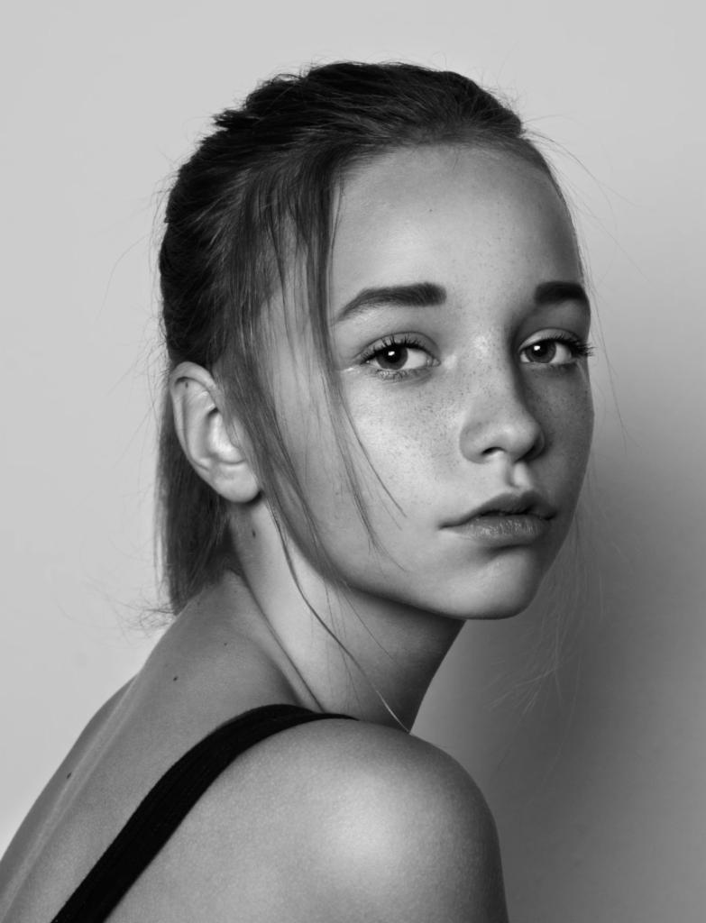 Portret młodej kobiety w czarno-białej tonacji, z delikatnymi piegami i włosami zaczesanymi do tyłu, spoglądającej przez ramię