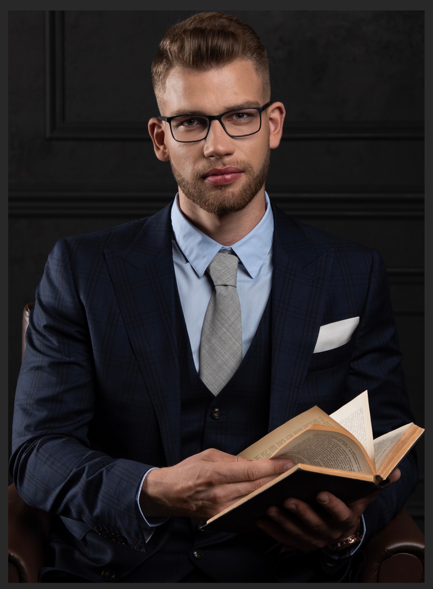 Mężczyzna w garniturze, okularach i z książką, pozujący na tle ciemnej ściany z dekoracyjnymi panelami.