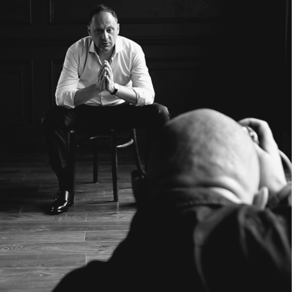 Czarno-białe zdjęcie zza kulis sesji biznesowej: mężczyzna siedzi na krześle z rękami złożonymi jak do modlitwy, patrzy w obiektyw, w tle ciemne panele ścienne, a na pierwszym planie głowa...