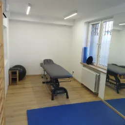 Jasne wnętrze gabinetu z drabinką gimnastyczną, szarym stołem do masażu, piłką rehabilitacyjną na stołku i niebieską matą podłogową.