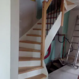 Drewniane schody z białymi podstopnicami i jasnymi drewnianymi stopniami w trakcie montażu, widoczna drabina i fragment instalacji na ścianie w surowym wnętrzu.