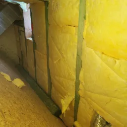 Żółta wełna mineralna w przestrzeni poddasza, zamontowana między zielonymi drewnianymi belkami, widoczny fragment wentylacji aluminiowej.