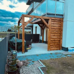 Drewniana pergola przy balkonie nowoczesnego budynku, widok z ogrodu z kostką brukową, elementami małej architektury i widocznym fragmentem elewacji.