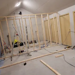 Stężyca: Konstrukcja szkieletowa z drewnianych belek tworząca ścianę działową w pomieszczeniu z widocznymi narzędziami i materiałami budowlanymi, przygotowana do montażu płyty karton gips.