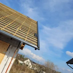 Konstrukcja drewniana dachu z widocznymi łatami i kontrłatami, przygotowana pod pokrycie dachowe, z zamontowaną rynną, na tle błękitnego nieba.