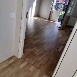 Jasny parkiet ułożony w jodełkę w pokoju z otwartymi drzwiami balkonowymi, widok na ogród i domy w tle.