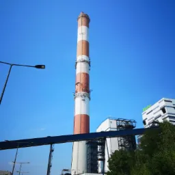 Wysoki komin przemysłowy z czerwono-białymi pasami na tle błękitnego nieba, widoczny fragment konstrukcji stalowej i budynek biurowy z zielonym logo.