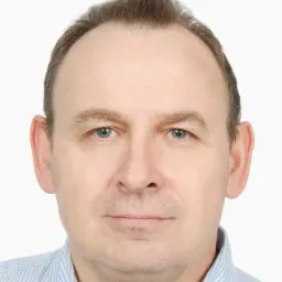 Portret mężczyzny w niebieskiej koszuli w paski, z delikatnym uśmiechem i jasnymi oczami, na białym tle.