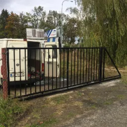 Czarna, metalowa brama wjazdowa, częściowo zamontowana, leżąca na ziemi obok otwartego busa na tle drzew i billboardów.