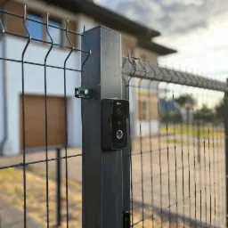 Czarny wideodomofon marki Dahua zamontowany na słupku ogrodzenia panelowego z widokiem na nowoczesny dom jednorodzinny w tle.