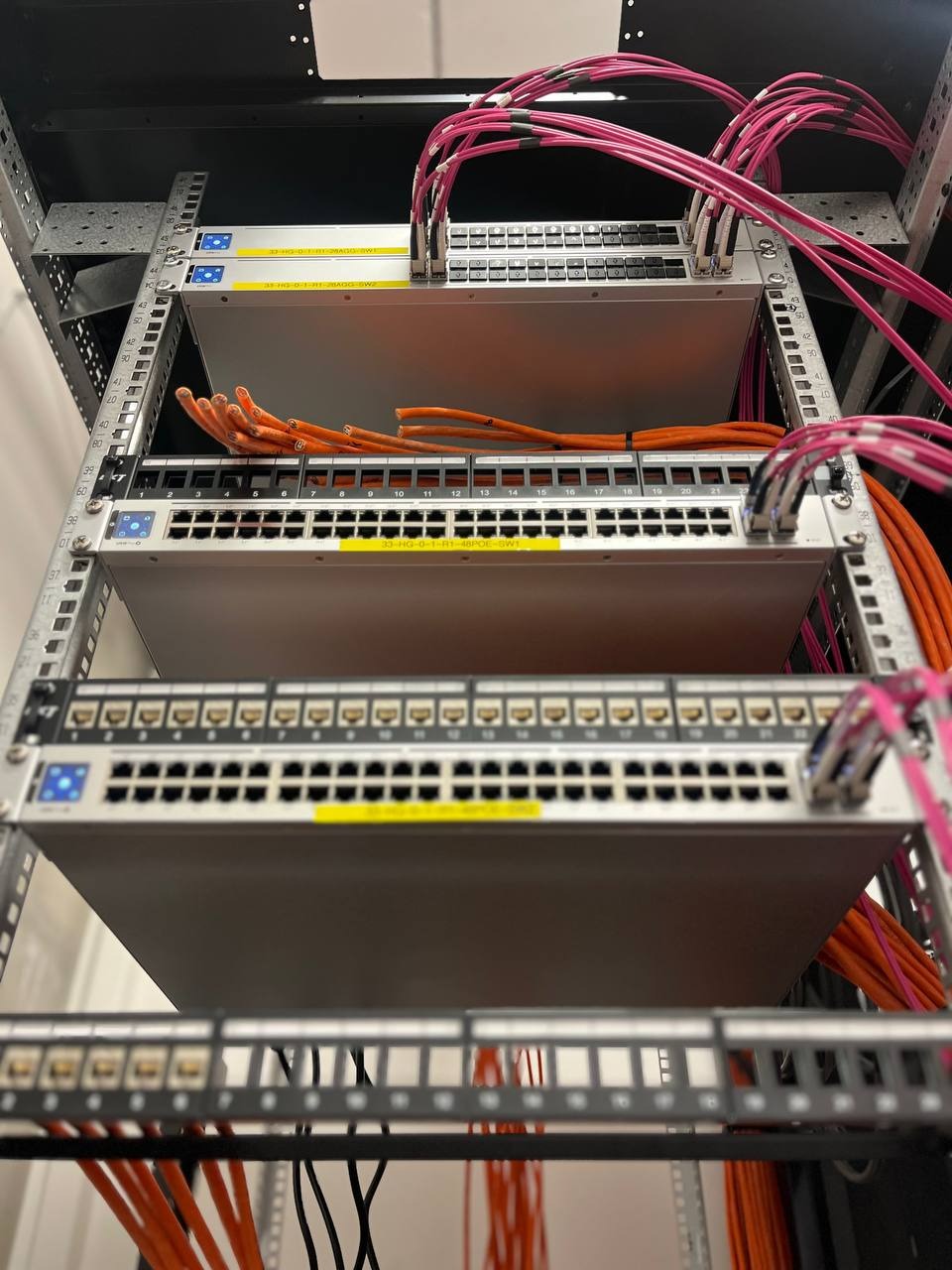 Widok z dołu na zainstalowane switche sieciowe w szafie rack we Wrocławiu, z widocznym okablowaniem strukturalnym w kolorach pomarańczowym i różowym.