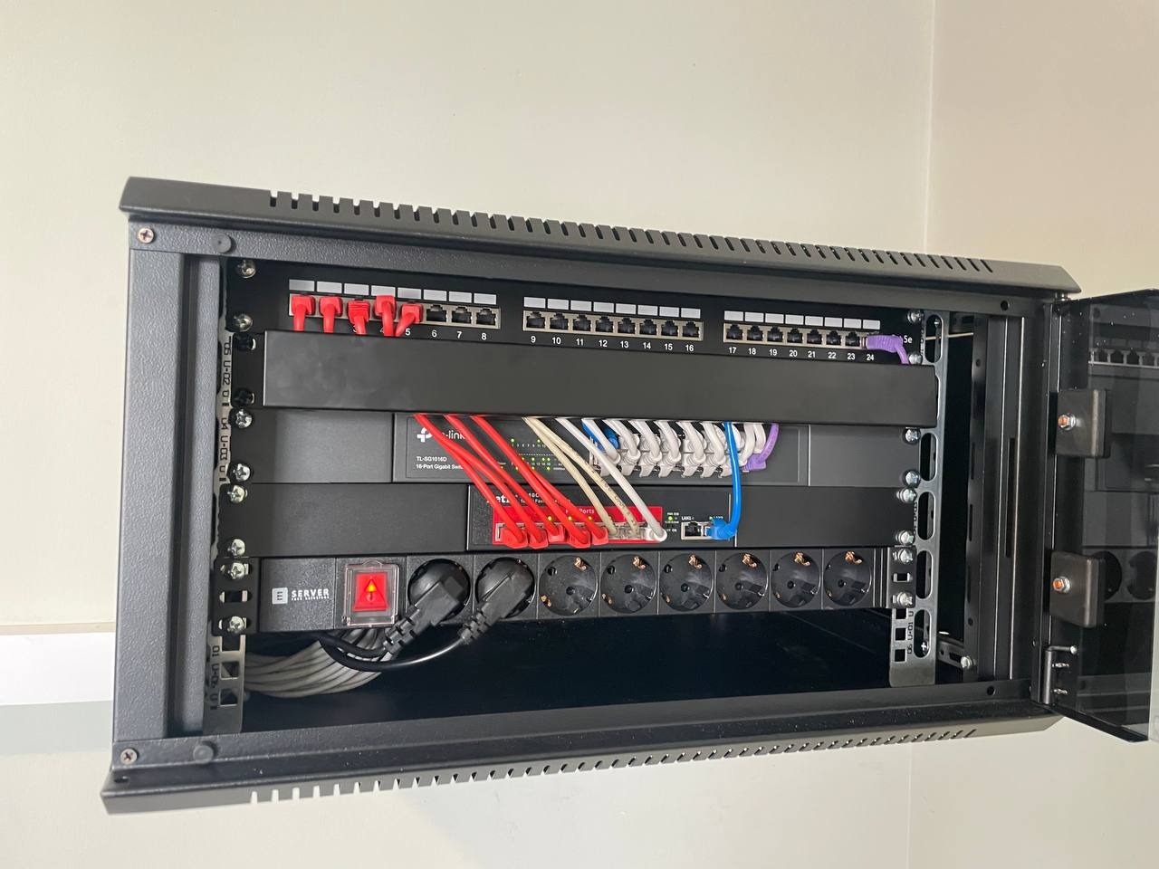 Otwarta szafa rack z zamontowanym panelem krosowniczym, switchem Gigabit Ethernet, listwą zasilającą z czerwonym włącznikiem i uporządkowanym okablowaniem sieciowym.