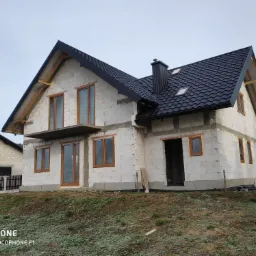 Surowy stan dwukondygnacyjnego domu z białych bloczków, z zamontowanymi oknami z brązowymi ramami i czarnym dachem z blachodachówki, widoczne elementy więźby dachowej, w tle podobny dom i zalesione...