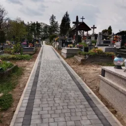 Cmentarna alejka z szarej kostki brukowej z ciemnoszarym pasem pośrodku, prowadząca w kierunku nagrobków i kapliczki pod pochmurnym niebem.