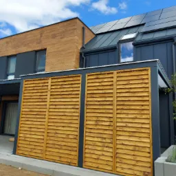 Nowoczesny dom z panelami słonecznymi na dachu, drewnianą elewacją i drewnianą przesłoną oddzielającą taras, z widocznymi elementami zieleni w betonowych donicach.