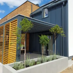 Nowoczesny dom z zadaszonym tarasem i drewnianymi panelami, ozdobiony podłużnymi donicami z lawendą i drzewkami, widoczne narzędzia budowlane.