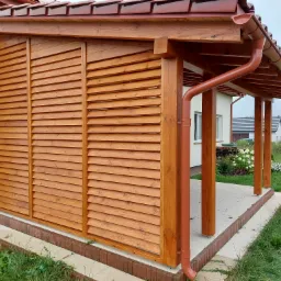 Zadaszona konstrukcja z drewnianymi żaluzjami i brązowymi rynnami, osłaniająca taras przy domu z widocznym ogrodem w tle.