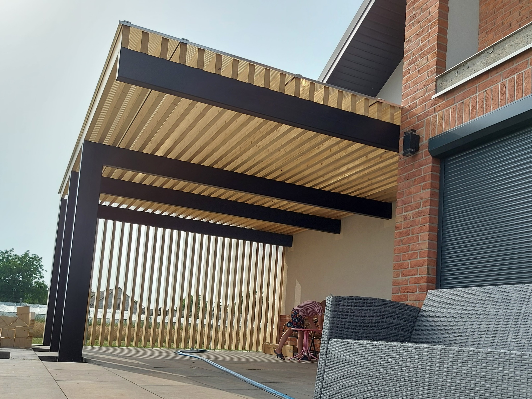 Nowoczesna pergola z drewnianym ażurem i czarną ramą na tle ceglanego domu z roletami, z widocznym fragmentem mebli ogrodowych i osobą pracującą wewnątrz konstrukcji.
