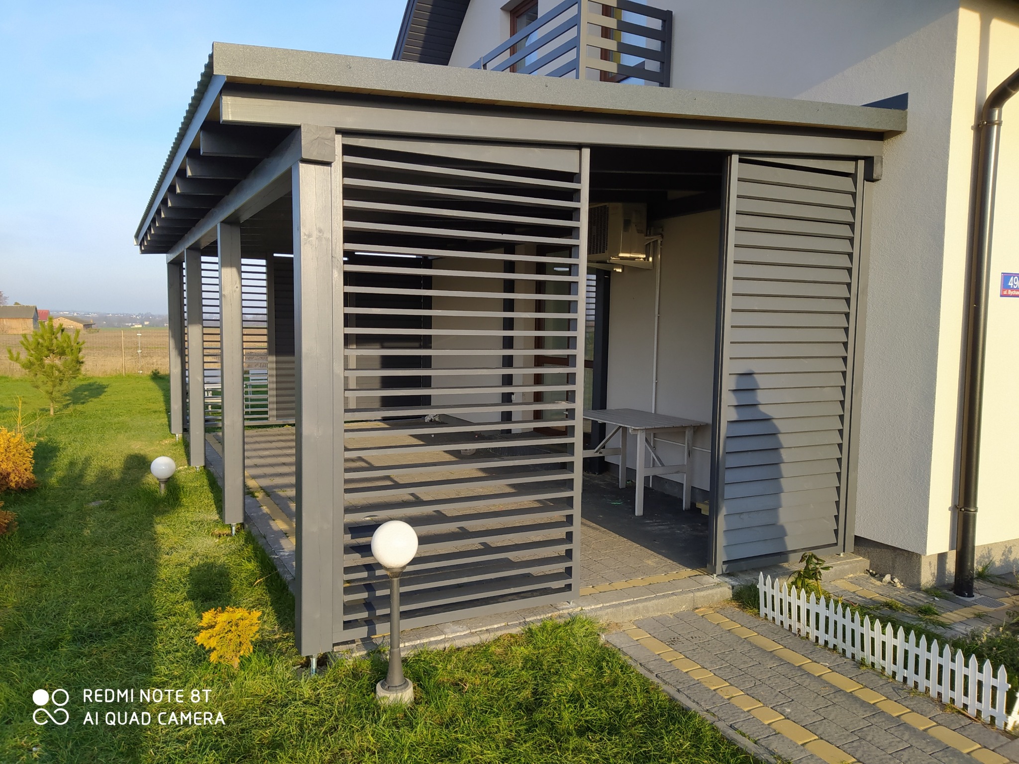 Szara konstrukcja z poziomych listew, częściowo zabudowana, przylegająca do budynku z białymi ścianami, oświetlona kulistymi lampami ogrodowymi.