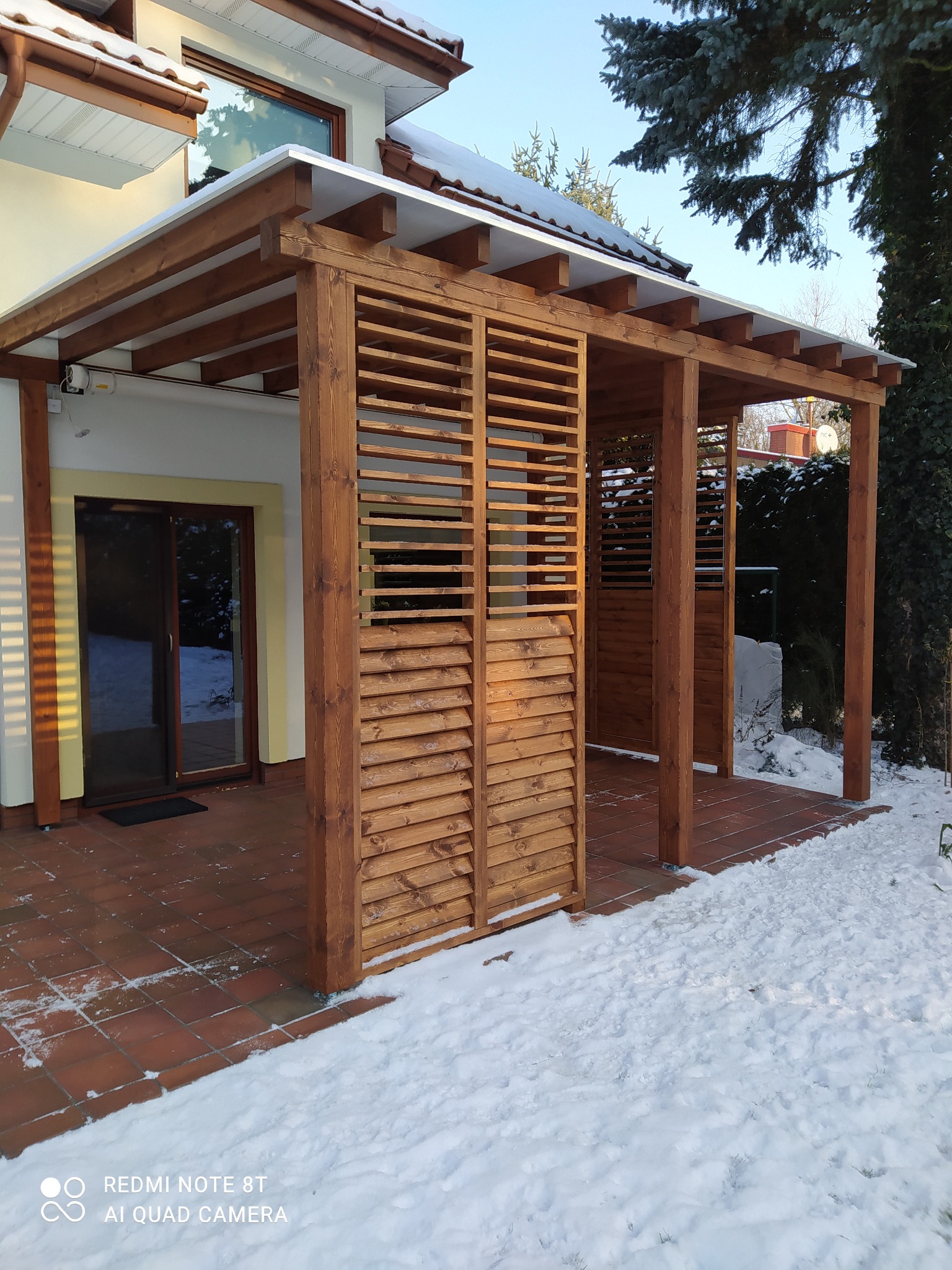 Drewniana konstrukcja z lamelami osłaniająca taras z brązowej terakoty, widok zimowy z fragmentami śniegu, zadaszenie z prześwitującym poliwęglanem.