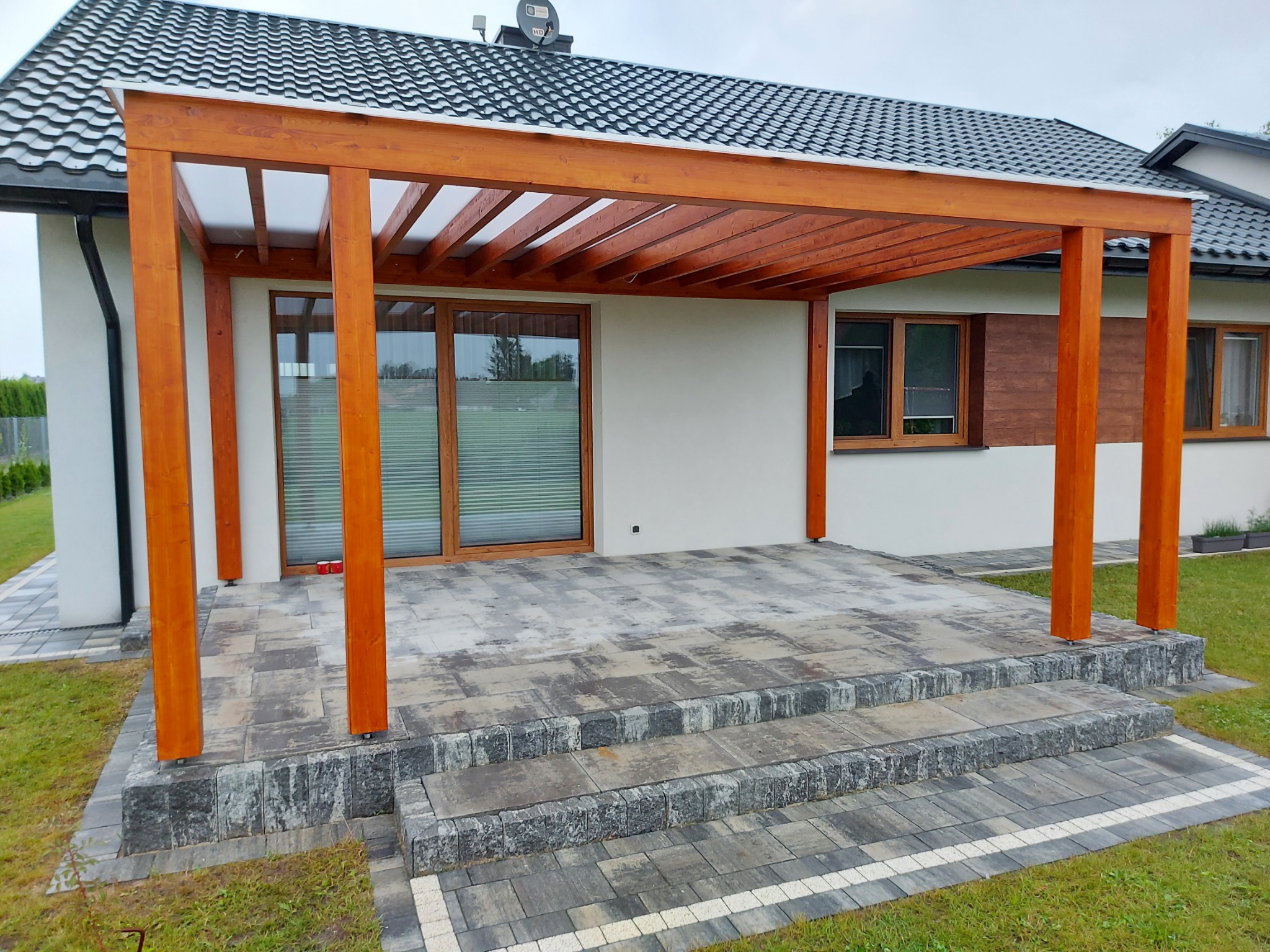 Drewniana konstrukcja z zadaszeniem nad tarasem z kostki brukowej, widok na dom z szarą dachówką.