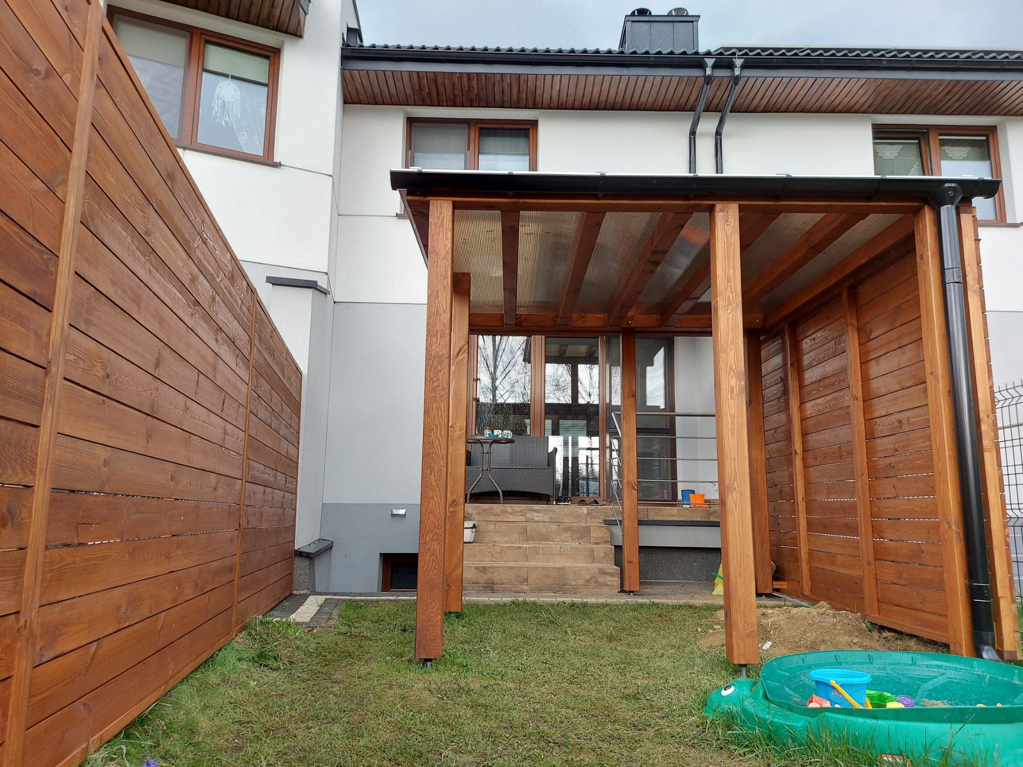 Zadaszenie z drewna nad tarasem z poliwęglanowym dachem, widok z ogrodu, obok drewniane ogrodzenie i piaskownica w kształcie żółwia.