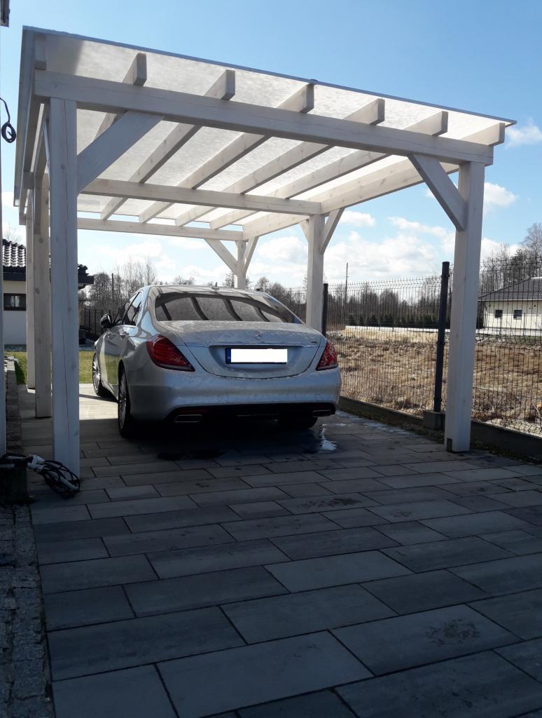 Biała drewniana konstrukcja z zadaszeniem nad miejscem parkingowym, pod którą stoi srebrny samochód osobowy na szarej kostce brukowej. Widoczne ogrodzenie i zabudowa jednorodzinna w tle.