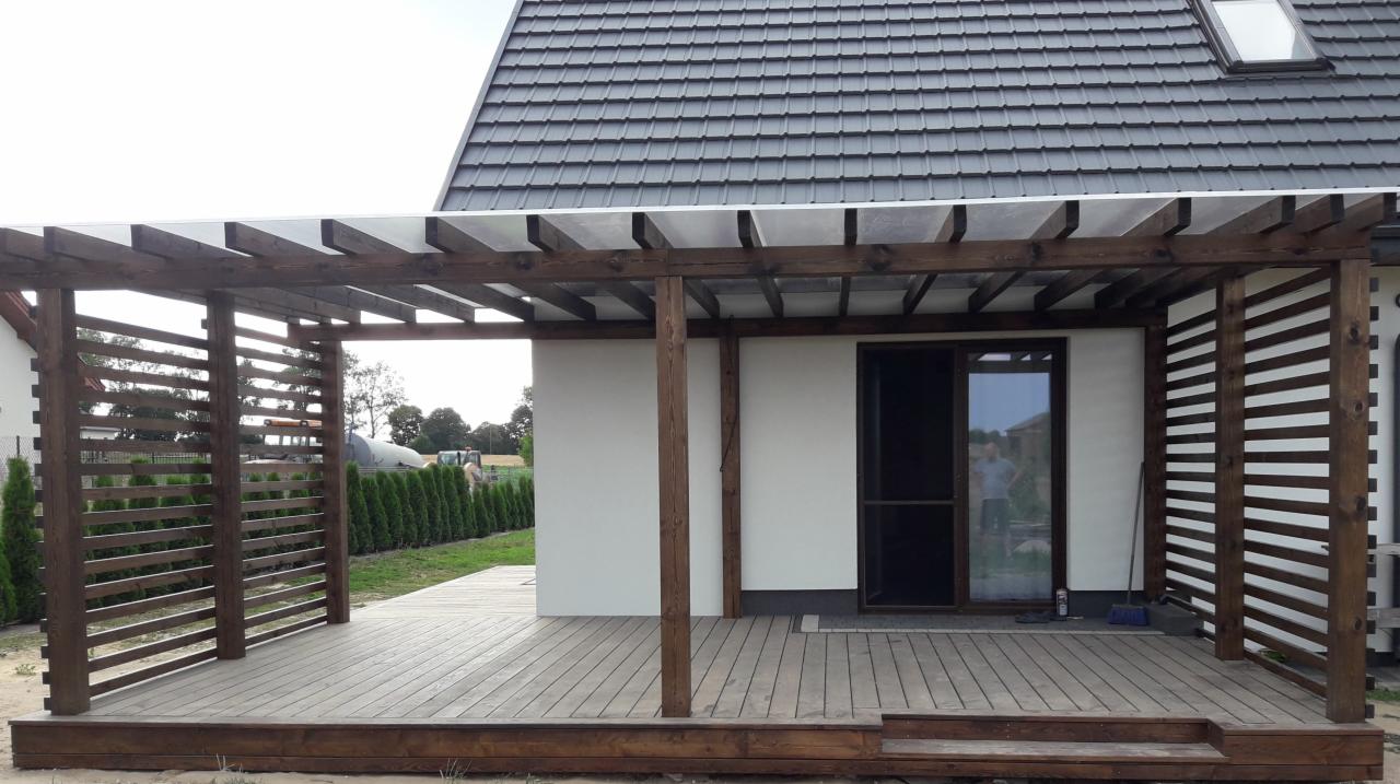 Zadaszona drewniana konstrukcja z ażurowymi ścianami i drewnianym tarasem przylegająca do białego budynku z szarym dachem, widoczne tuje w tle.