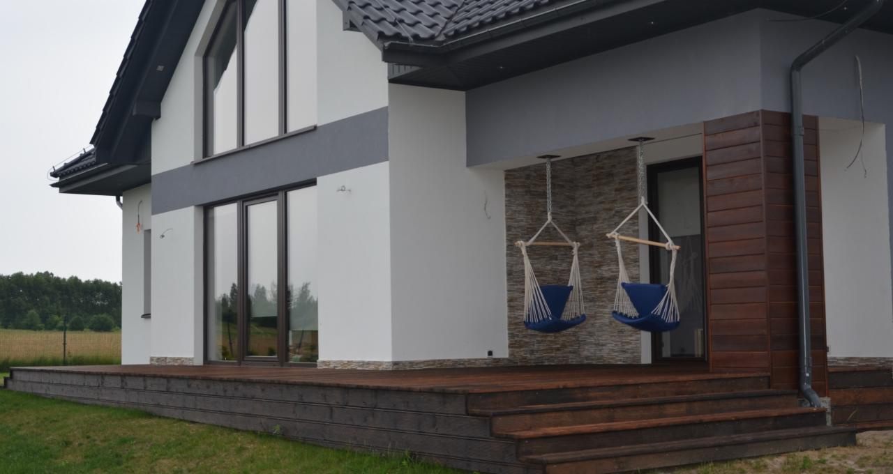 Nowoczesny dom z drewnianym tarasem i dwoma wiszącymi fotelami hamakowymi w kolorze niebieskim, widoczne schody tarasowe i okładzina ściany imitująca kamień.