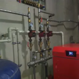 Czerwony piec centralnego ogrzewania z podłączonymi rurami, manometrami i pompami obiegowymi w kotłowni o surowych ścianach.