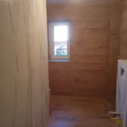 Wnętrze łazienki z drewnopodobnymi płytkami na ścianach i podłodze, widoczne okno z widokiem na zewnątrz oraz fragment zabudowy toalety.