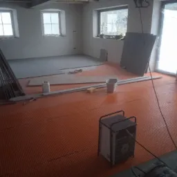 Wnętrze w trakcie remontu: pomarańczowa mata grzewcza pod płytki na podłodze, częściowo ułożone szare płytki, narzędzia glazurnicze, nagrzewnica powietrza i okna w tle