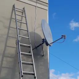Srebrna drabina oparta o ścianę budynku obok zainstalowanej satelitarnej anteny parabolicznej na tle błękitnego nieba z chmurami.