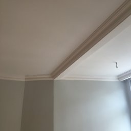 LAPIS HOME SP. Z O. O. - Pomieszczenie po odświeżeniu: szare ściany, biały sufit z ozdobnymi listwami i wystającym kablem od lampy.