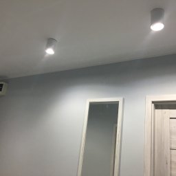 LAPIS HOME SP. Z O. O. - Pomieszczenie po malowaniu na szaro z trzema białymi lampami sufitowymi, białym lustrem i drzwiami z jasnego drewna.