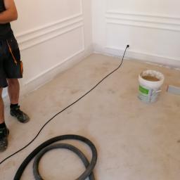 LAPIS HOME SP. Z O. O. - Przygotowanie do prac wykończeniowych: białe ściany z dekoracyjnymi listwami, podłoga po wylewce, wiadro z zaprawą i wąż odkurzacza przemysłowego.