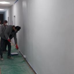 LAPIS HOME SP. Z O. O. - Mężczyzna w masce maluje na biało ścianę w korytarzu biurowym za pomocą wałka, zabezpieczona taśmą malarską, podłoga przykryta folią ochronną.