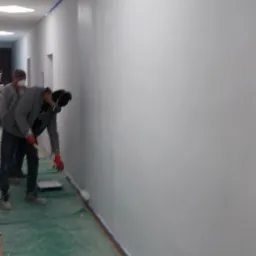 Mężczyzna w masce maluje na biało ścianę w korytarzu biurowym za pomocą wałka, zabezpieczona taśmą malarską, podłoga przykryta folią ochronną.
