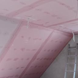 LAPIS HOME SP. Z O. O. - Montaż różowych płyt kartonowo-gipsowych na poddaszu ze skosami, widoczne kable elektryczne, drabina aluminiowa i okno dachowe zabezpieczone folią.