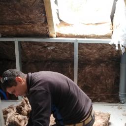 LAPIS HOME SP. Z O. O. - Pracownik w okularach ochronnych montuje izolację z wełny mineralnej w poddaszu, widoczne rury kanalizacyjne, stelaż podtynkowy Geberit i drabina aluminiowa.