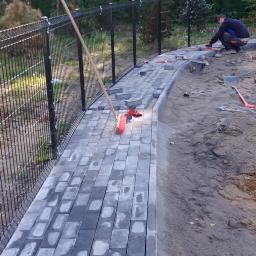 LAPIS HOME SP. Z O. O. - Układanie szarej kostki brukowej na posesji w Gdańsku, widoczny pracownik, narzędzia i częściowo ułożony chodnik wzdłuż ogrodzenia.